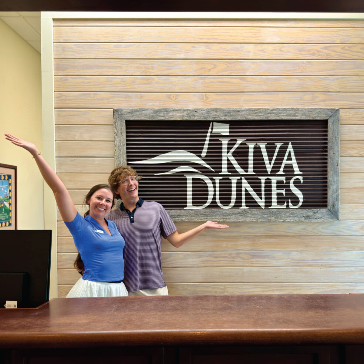 Two smiling staff members stand behind a wooden reception desk with arms raised in a friendly gesture in front of a large wall sign reading 'Kiva Dunes'.